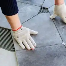Handwerker mit Arbeitshandschuhen verlegt graue Bodenfliesen auf frischem Fliesenkleber