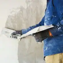 Handwerker trägt mit Glättkelle frischen Putz auf Innenwand auf, Arbeitskleidung mit Farbspritzern